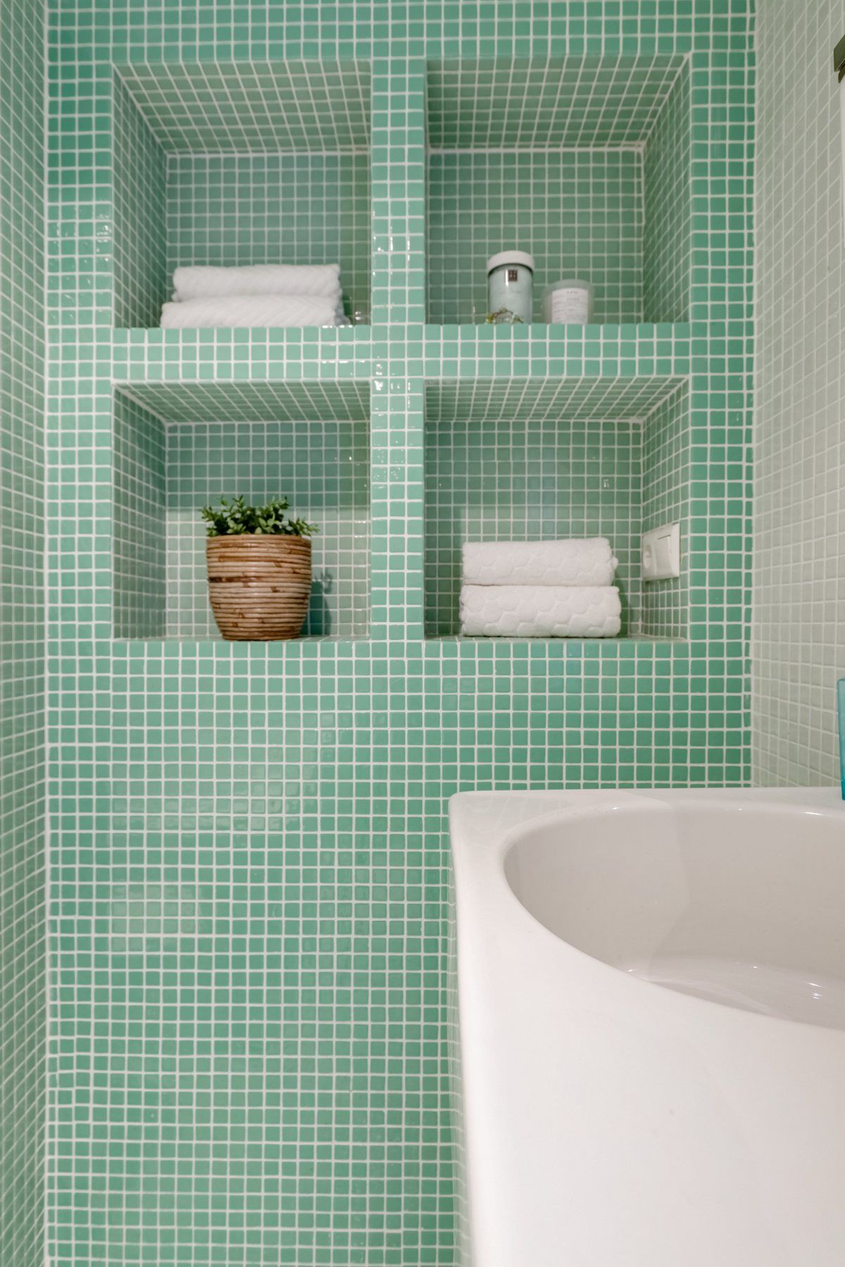 Salle de bain en mosaïque vert d'eau avec niches maçonnées, serviettes blanches et pot en rotin